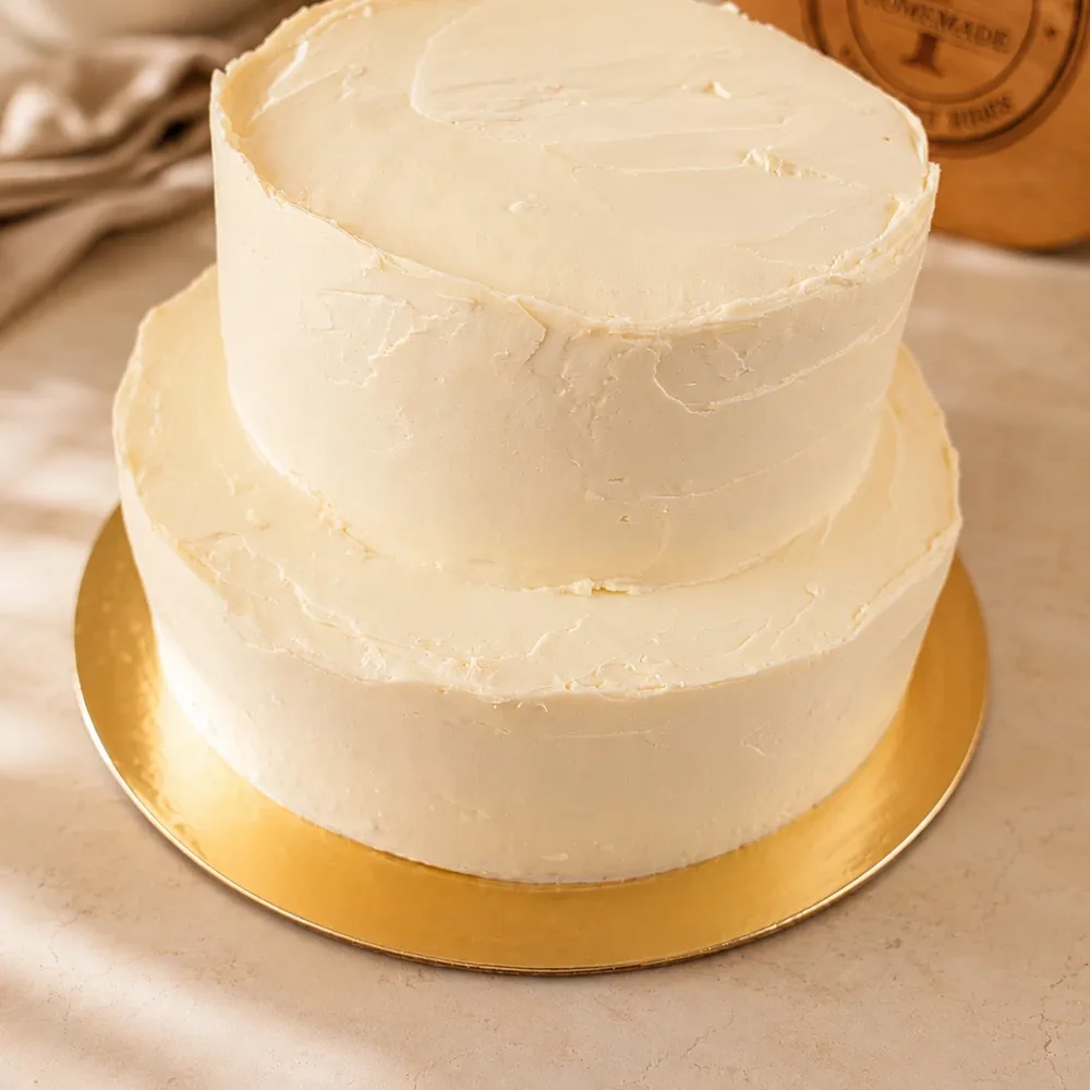 A two tier vanilla cake finished in smooth pale cream cheese frosting on a gold board.